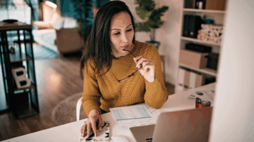 Woman sitting at her desk in her living room at home holding her eyeglasses in her hand while working on her laptop