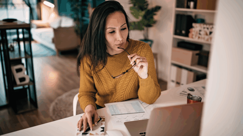 Woman sitting at her desk in her living room at home holding her eyeglasses in her hand while working on her laptop